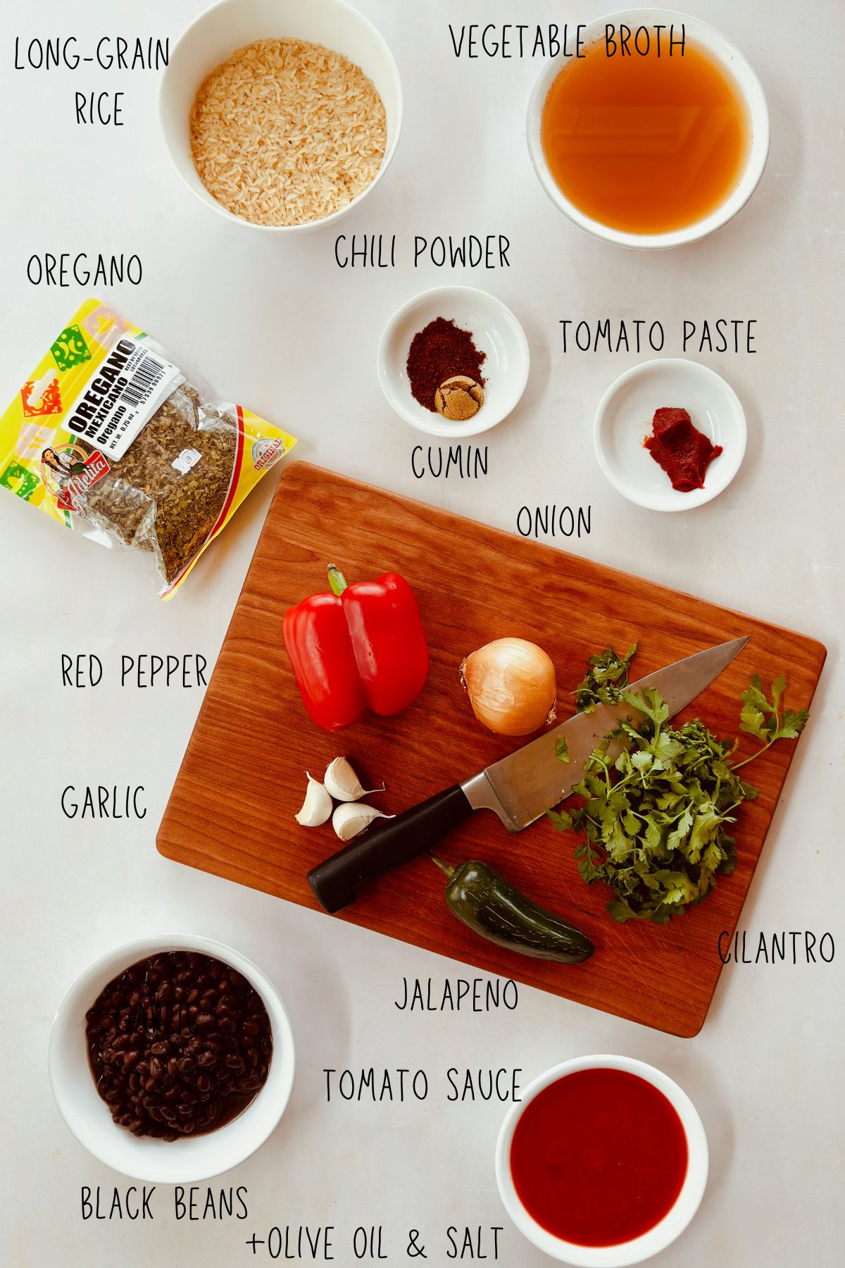 Ingredients for Mexican-Style Black Beans and Rice including long-grain rice, black beans, red pepper, jalapeño, cilantro, garlic, onion, tomato sauce, tomato paste, vegetable broth, chili powder, cumin, and oregano arranged on a white surface with a wooden cutting board.