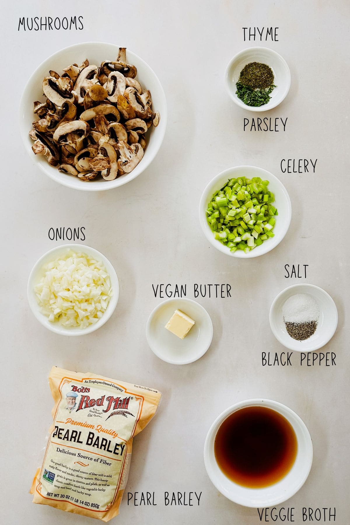 Ingredients for mushroom barley pilaf arranged in small bowls on a white surface, including sliced mushrooms, chopped onions, chopped celery, vegan butter, thyme, parsley, salt, black pepper, pearl barley, and vegetable broth.