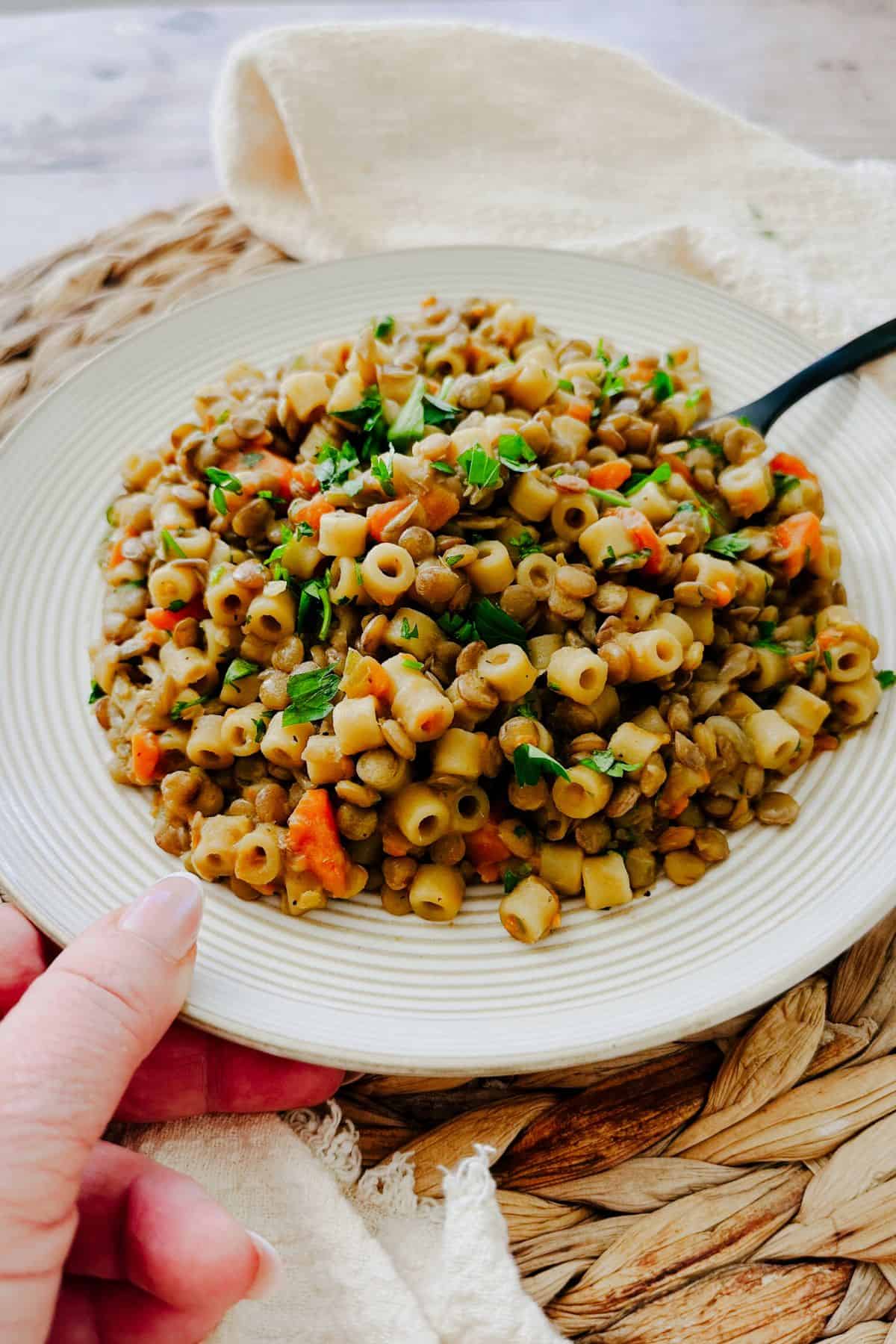Plate of one-pot lentils and ditalini pasta with carrots and herbs, held in hand over a woven placemat for a cozy, casual meal.