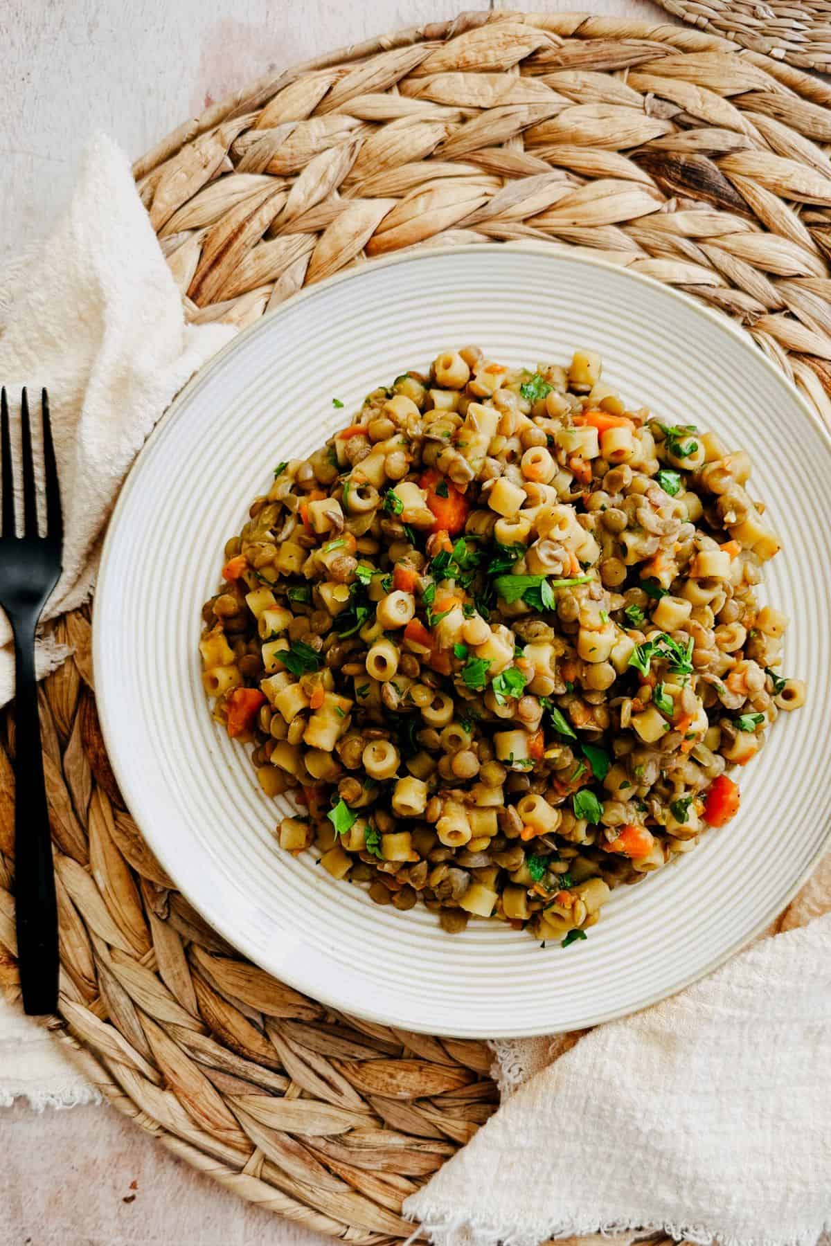 Overhead view of one-pot lentils and ditalini pasta with vegetables and parsley, served on a white plate with fork and linen nearby.