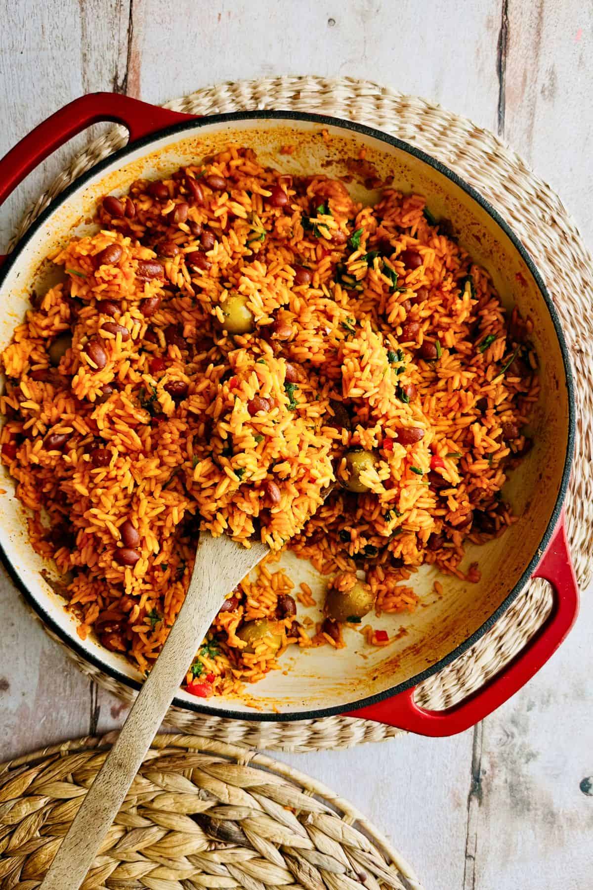 Overhead view of Puerto Rican yellow rice and beans with green olives in a red Dutch oven, with a wooden spoon in the pan.