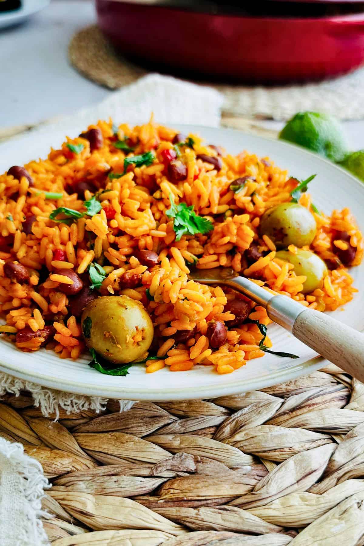 Plate of Puerto Rican yellow rice and beans with olives and cilantro, served on a woven placemat, with a fork resting in the rice.