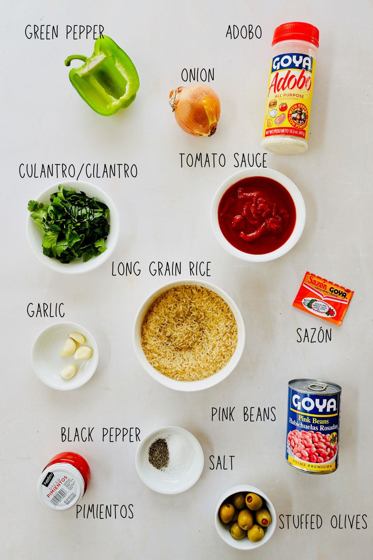 Flat lay of ingredients for Puerto Rican yellow rice and beans: rice, pink beans, tomato sauce, green pepper, onion, garlic, olives, sazón, adobo, and herbs.