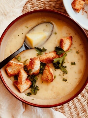 Roasted Garlic and Potato soup in a bowl topped with croutons and chopped parsley. Spoon is in the soup with potato and soup on it.