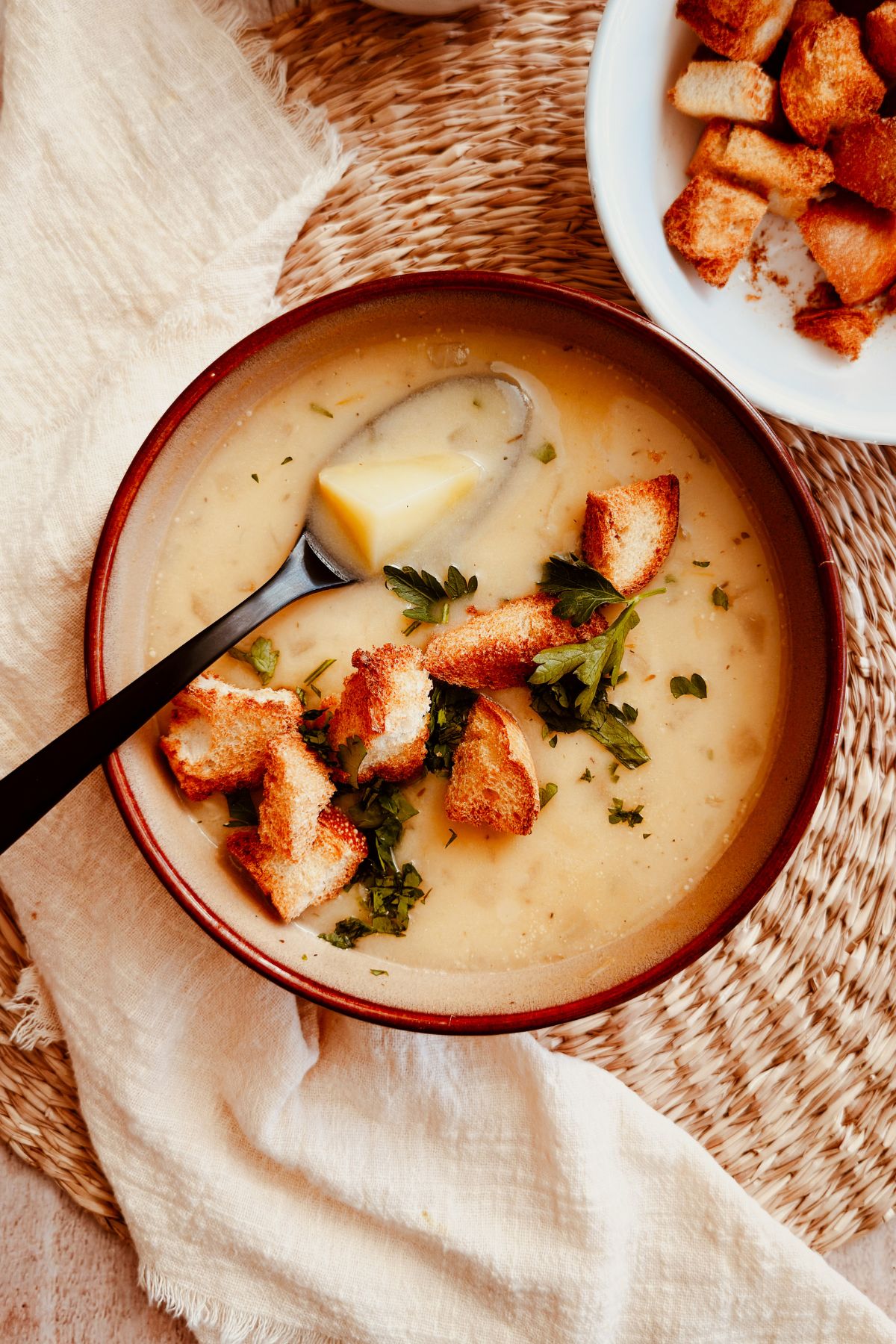 Creamy roasted garlic and potato soup served in a brown bowl, topped with golden croutons and fresh parsley, with a spoon lifting a tender chunk of potato. A small bowl of extra croutons sits nearby on a woven placemat with a light linen napkin.