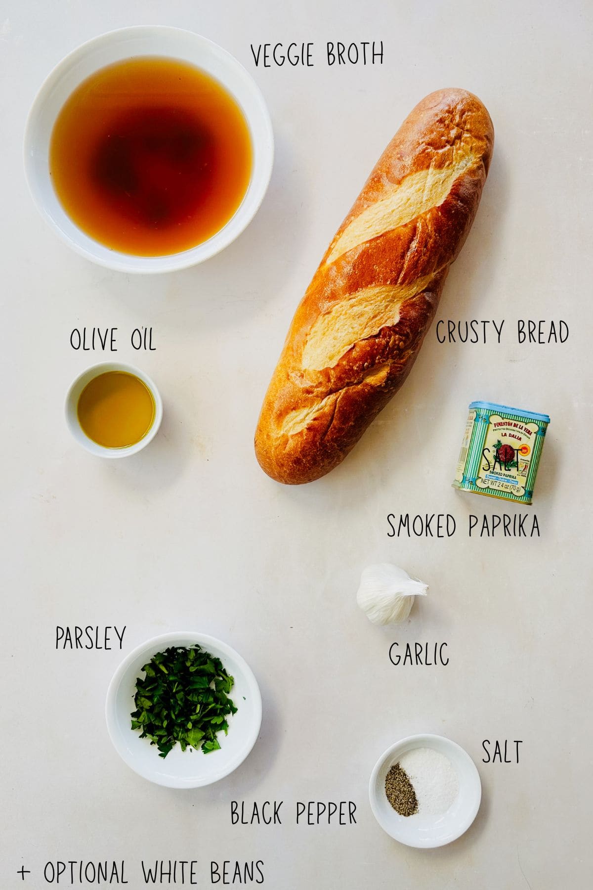 Labeled ingredients for Spanish garlic soup (sopa de ajo) including vegetable broth, olive oil, crusty bread, smoked paprika, garlic, fresh parsley, salt, black pepper, and optional white beans.