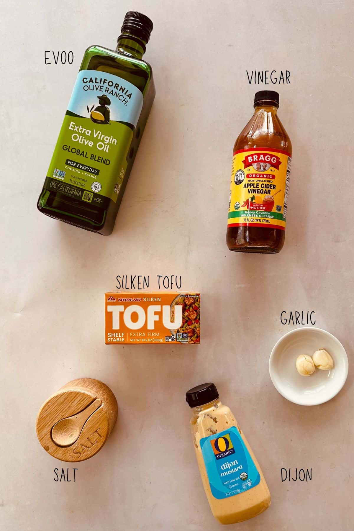 Labeled ingredients for tofu mayonnaise: olive oil, apple cider vinegar, silken tofu, garlic, salt, and dijon mustard arranged on white surface.