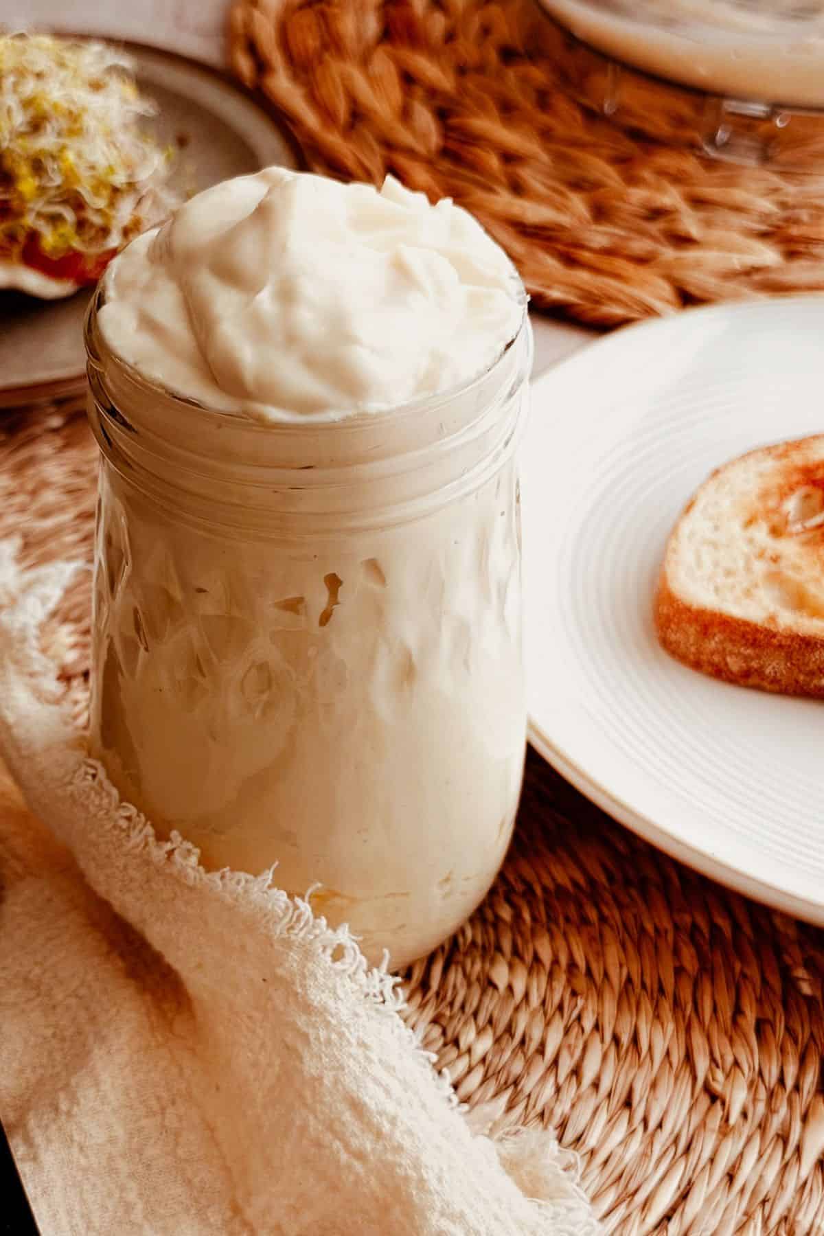 Jar of creamy vegan tofu mayo on a table setting with an open-faced sandwich in the back.