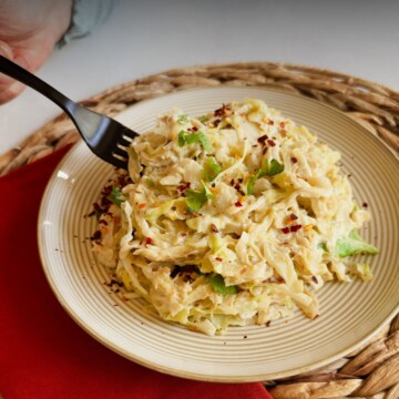 A black fork goes into a plate of creamy vegan cabbage alfredo topped with cilantro and red pepper flakes, with text reading 'How to Make Vegan Cabbage Alfredo - Delicious & Healthy.