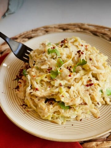 A black fork goes into a plate of creamy vegan cabbage alfredo topped with cilantro and red pepper flakes, with text reading 'How to Make Vegan Cabbage Alfredo - Delicious & Healthy.