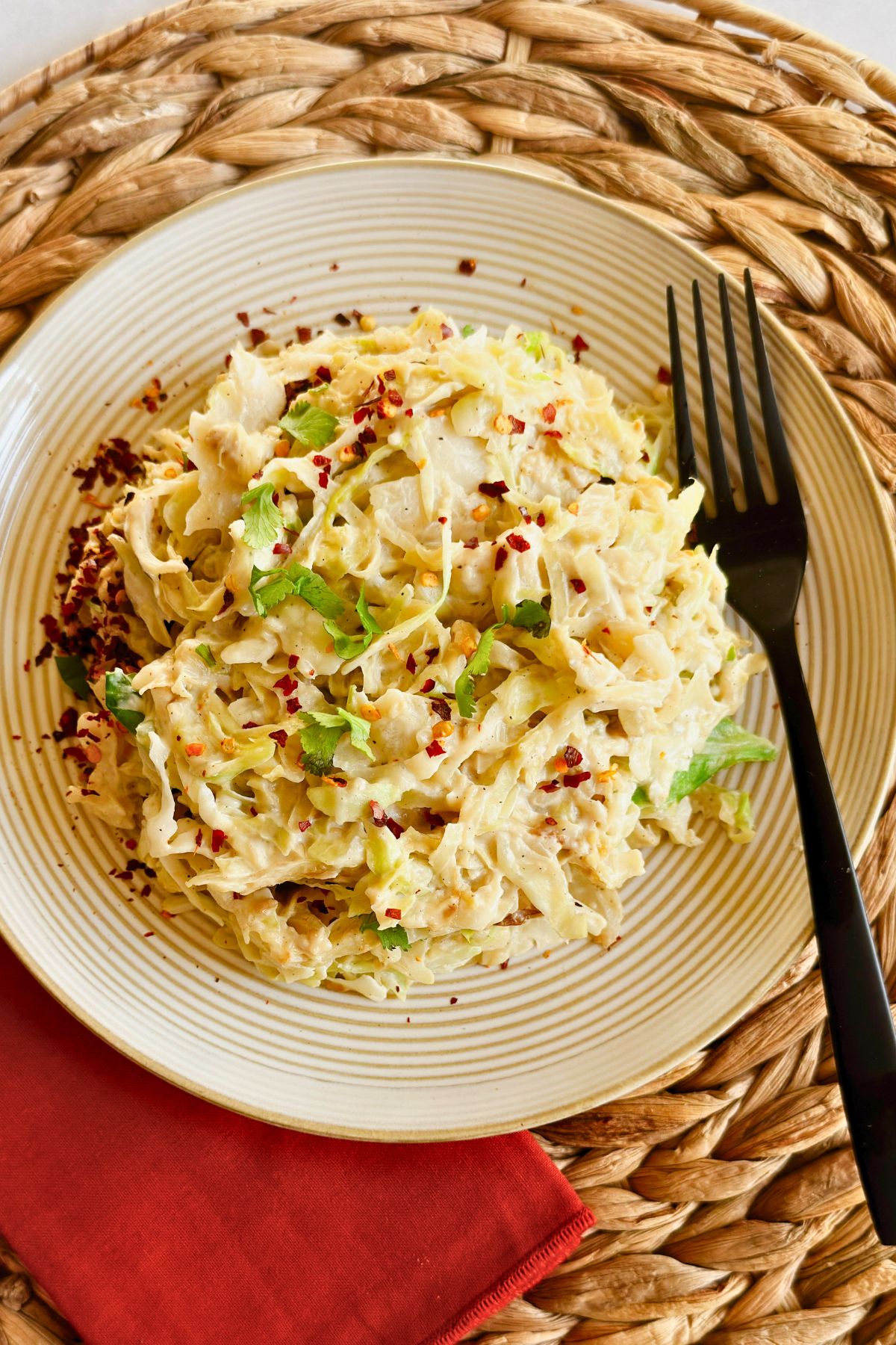 A plate of creamy vegan cabbage alfredo topped with fresh cilantro and red pepper flakes, served on a woven placemat with a black fork.