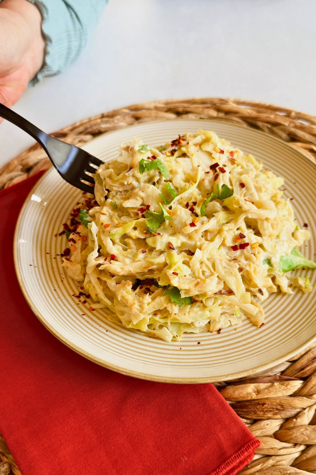 A hand holding a fork digging into a plate of creamy vegan cabbage alfredo topped with cilantro and red pepper flakes on a woven placemat.