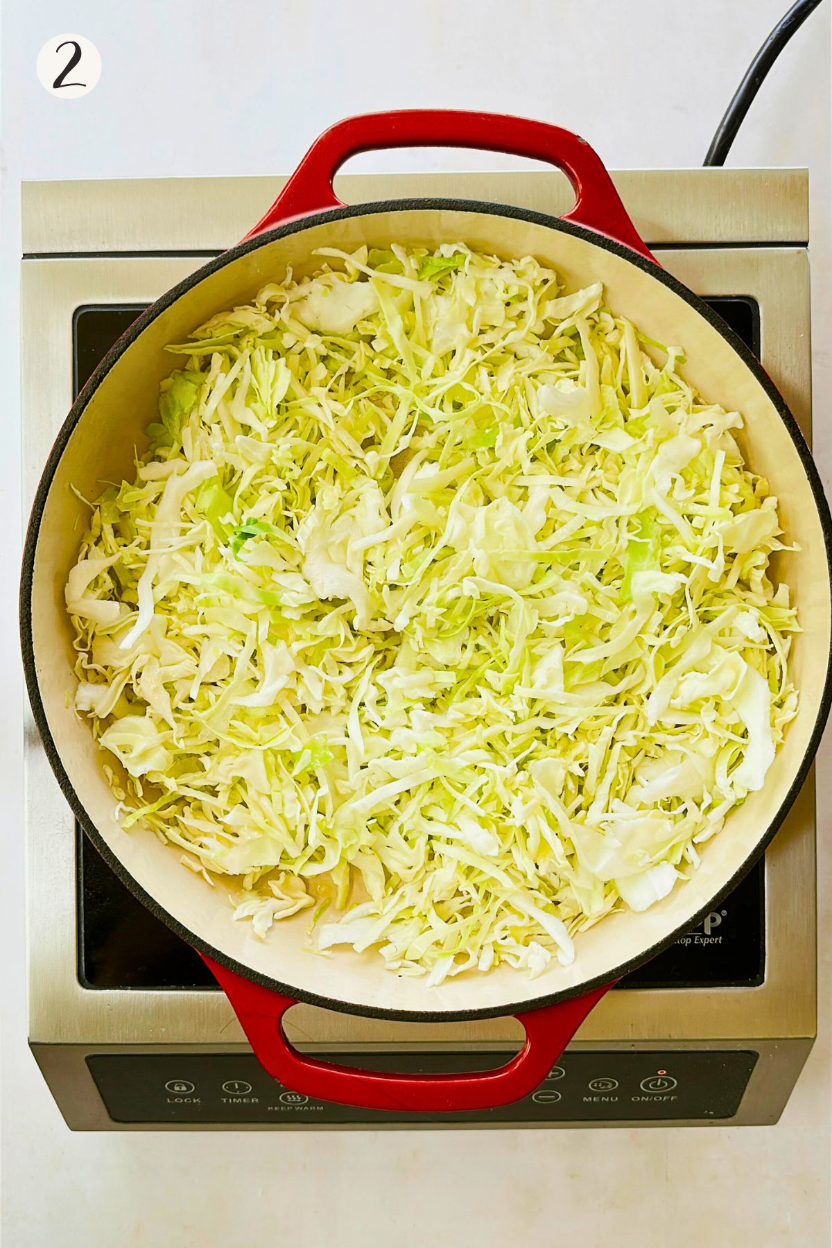 Shredded green cabbage sautéing in a red Dutch oven on an induction cooktop."
