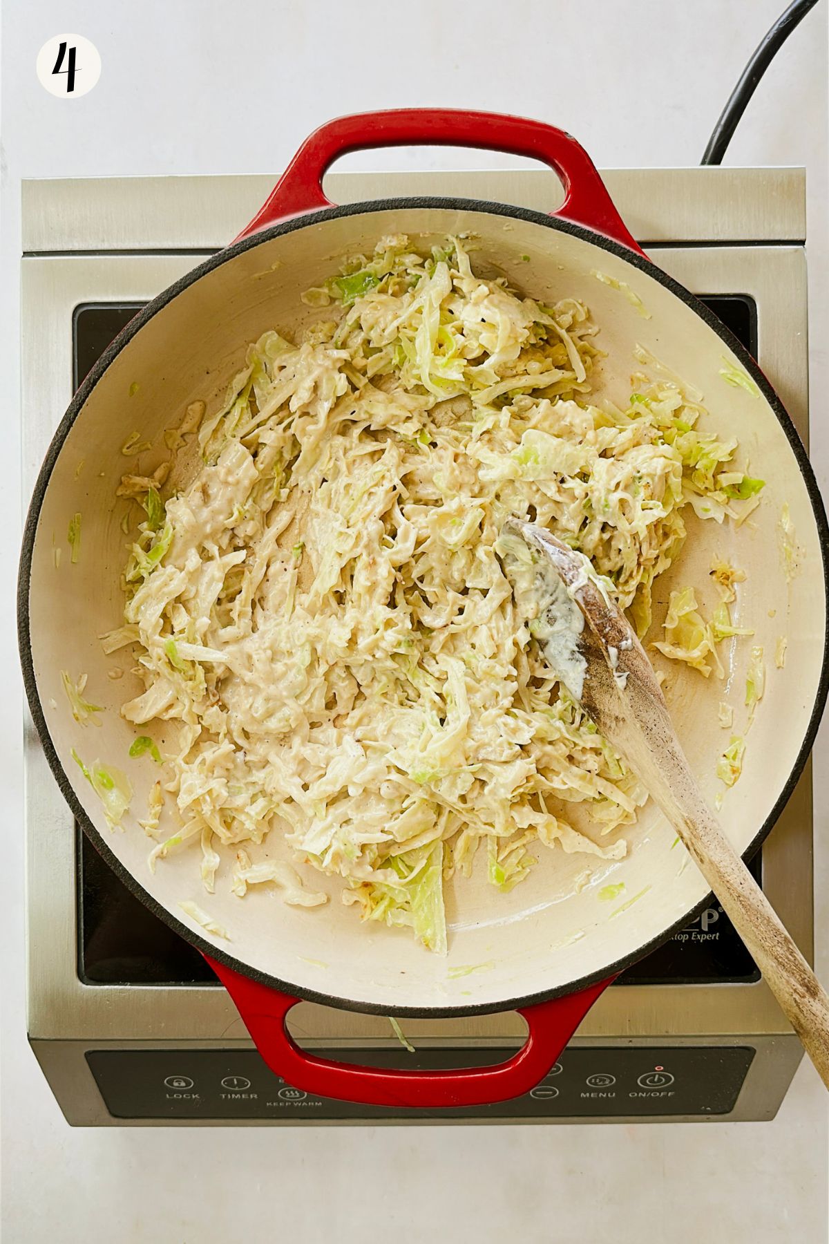 Cabbage and cashew alfredo sauce being stirred together in a red Dutch oven with a wooden spoon.