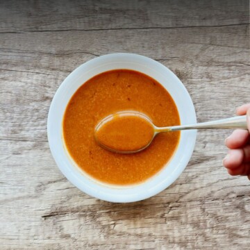 A hand holding a spoon over a white bowl filled with smooth, deep orange chipotle vinaigrette, set on a rustic wood surface.