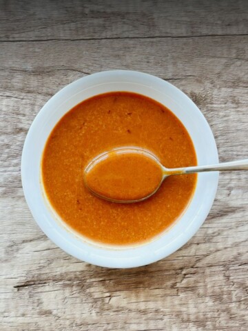 A hand holding a spoon over a white bowl filled with smooth, deep orange chipotle vinaigrette, set on a rustic wood surface.