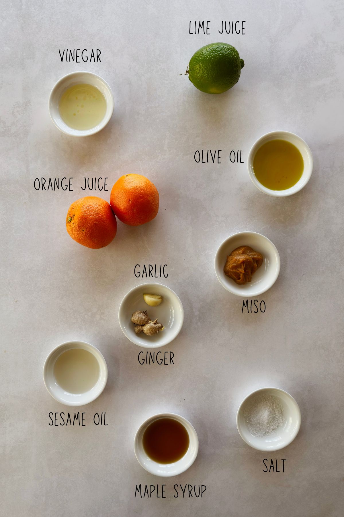 Flat lay of miso citrus dressing ingredients in small white bowls on a light gray surface, labeled: vinegar, lime juice, orange juice, olive oil, garlic, ginger, miso, sesame oil, maple syrup, and salt.
