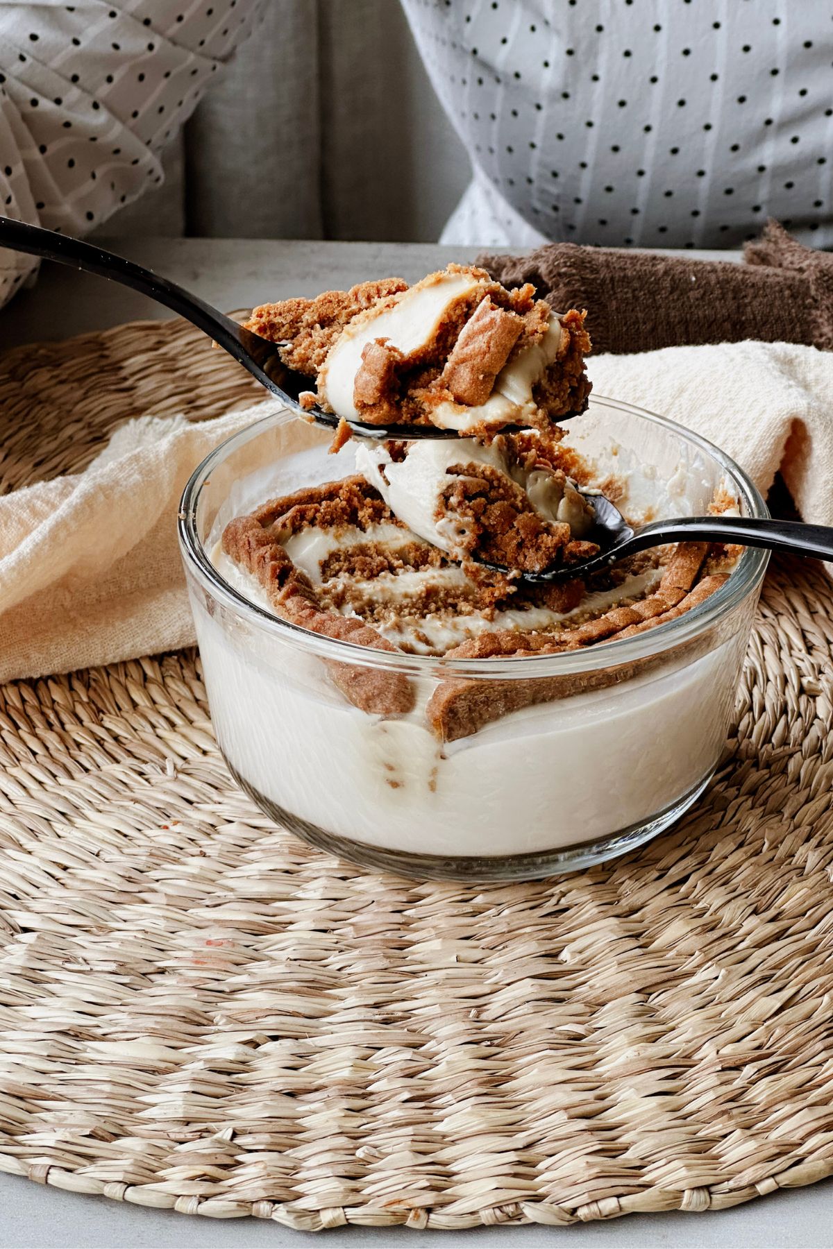 Two black spoons lifting a scoop of softened Biscoff cookies and creamy vegan Greek yogurt from a glass bowl on a woven placemat.