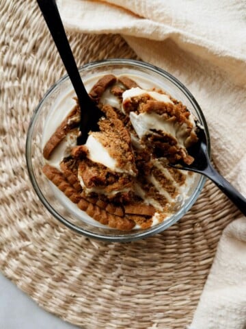 Overhead view of softened Biscoff cookies in a glass bowl of vegan Greek yogurt, being scooped with two black spoons on a woven placemat.