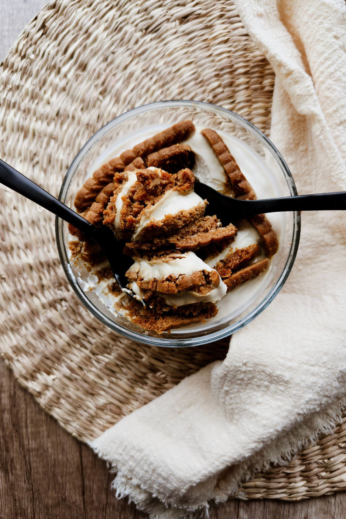 Overhead view of softened Biscoff cookies in a glass bowl of vegan Greek yogurt, being scooped with two black spoons on a woven placemat.