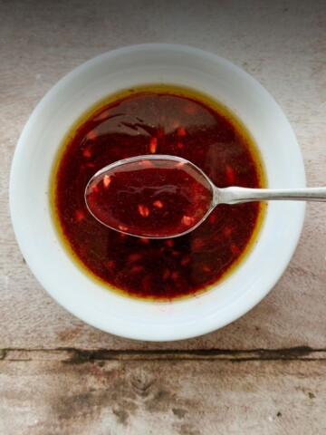 Close-up overhead shot of deep ruby pomegranate molasses vinaigrette in a white bowl with a spoon lifting the glossy dressing, on a rustic wood surface.