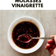 Overhead view of pomegranate molasses vinaigrette in a white bowl with a decorative spoon lifting the deep ruby dressing, with text overlay reading "How to Make Pomegranate Molasses Vinaigrette."