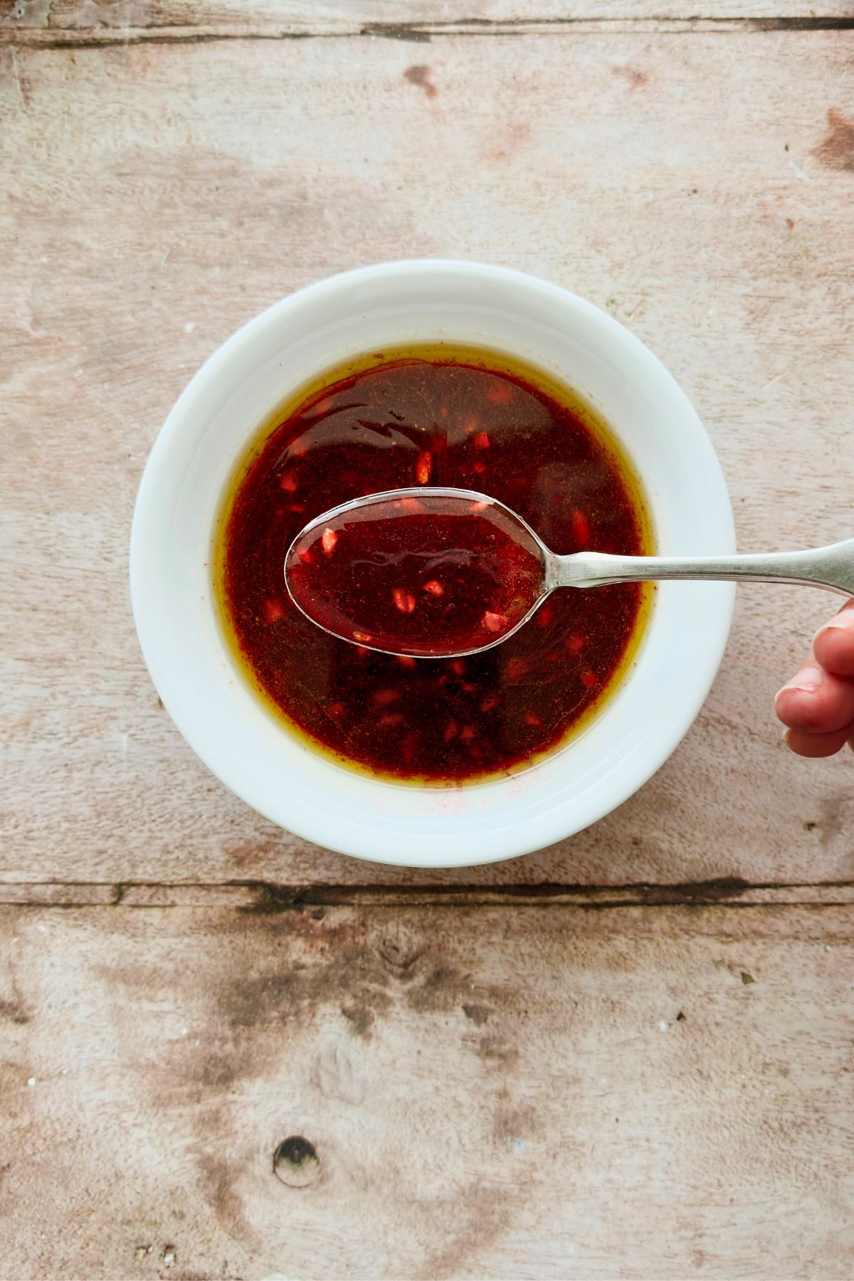 Close-up overhead shot of deep ruby pomegranate molasses vinaigrette in a white bowl with a spoon lifting the glossy dressing, on a rustic wood surface.