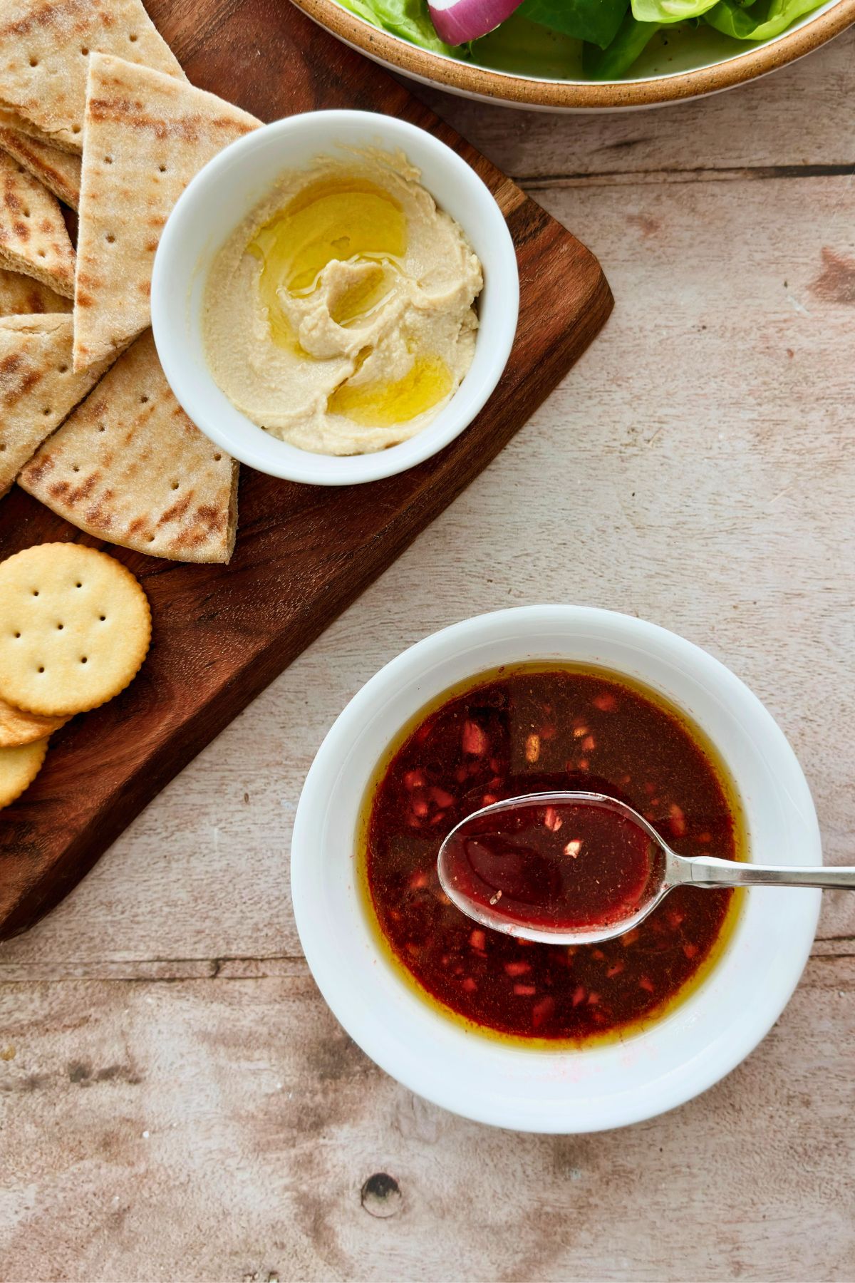 Overhead view of pomegranate molasses vinaigrette in a white bowl beside hummus drizzled with olive oil, pita, and crackers on a wooden board.