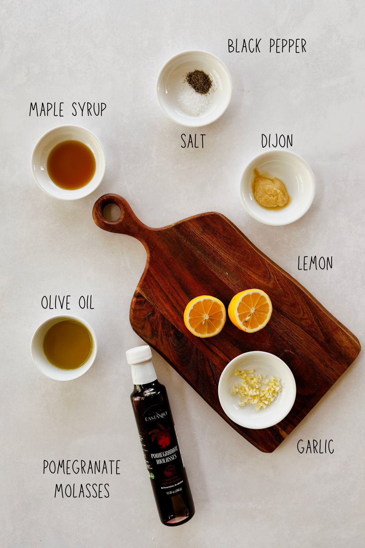Overhead flat lay of labeled pomegranate molasses vinaigrette ingredients including a bottle of pomegranate molasses, halved lemon, olive oil, maple syrup, Dijon, garlic, salt, and pepper.
