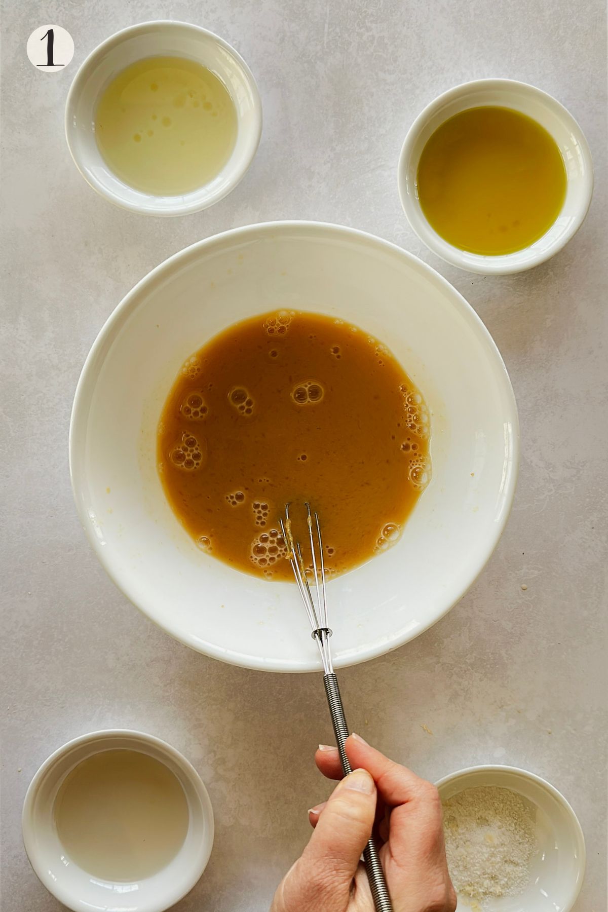 A hand whisks miso citrus dressing base in a large white bowl, surrounded by small bowls of vinegar, olive oil, sesame oil, and salt.