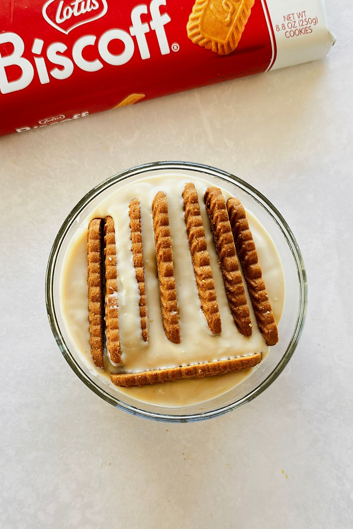 Biscoff cookies standing vertically in a glass bowl of thick vegan Greek yogurt, ready to refrigerate overnight.