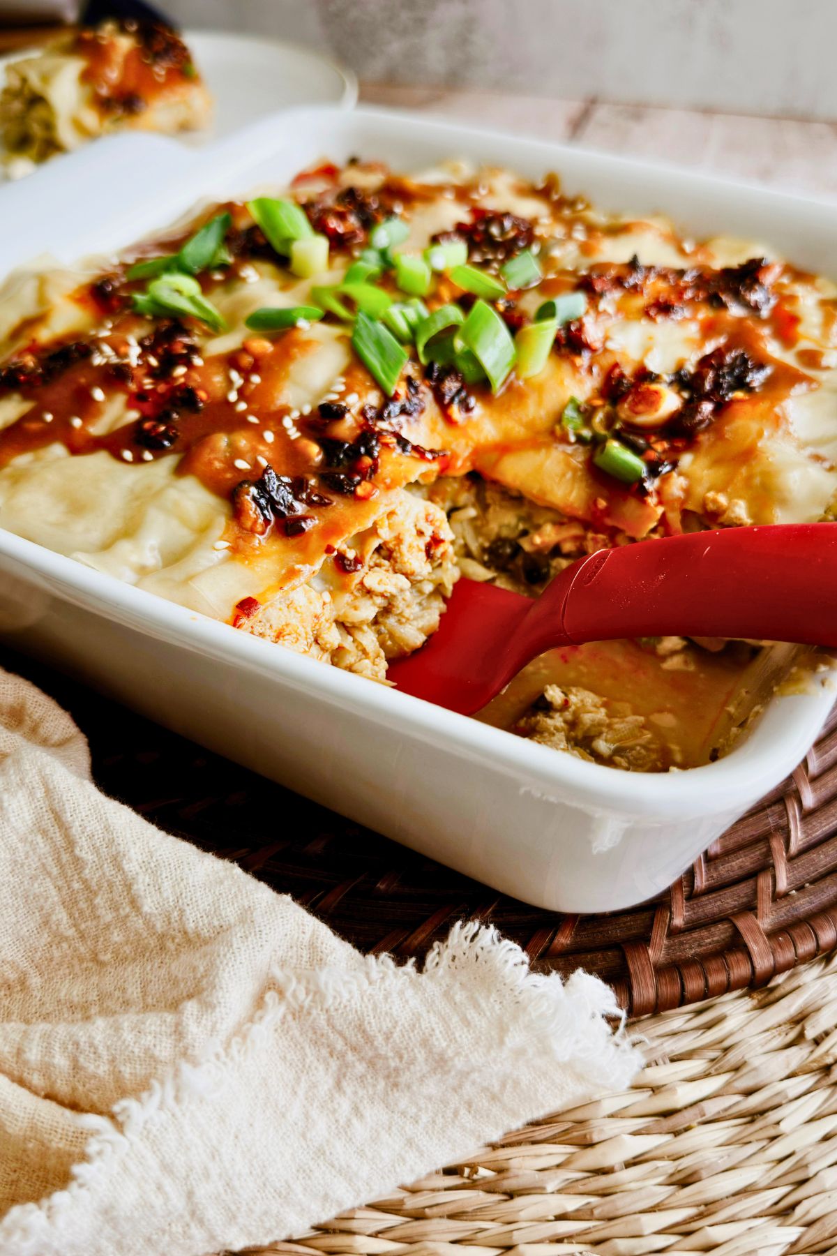 Vegan dumpling lasagna in a white baking dish, topped with crispy chili, sesame seeds, green onions, and peanut sauce, with a red spoon scooping out a serving.