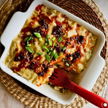 Overhead view of vegan dumpling lasagna with a serving removed, topped with crispy chili, sesame seeds, and green onions, with a red spatula resting in the dish.
