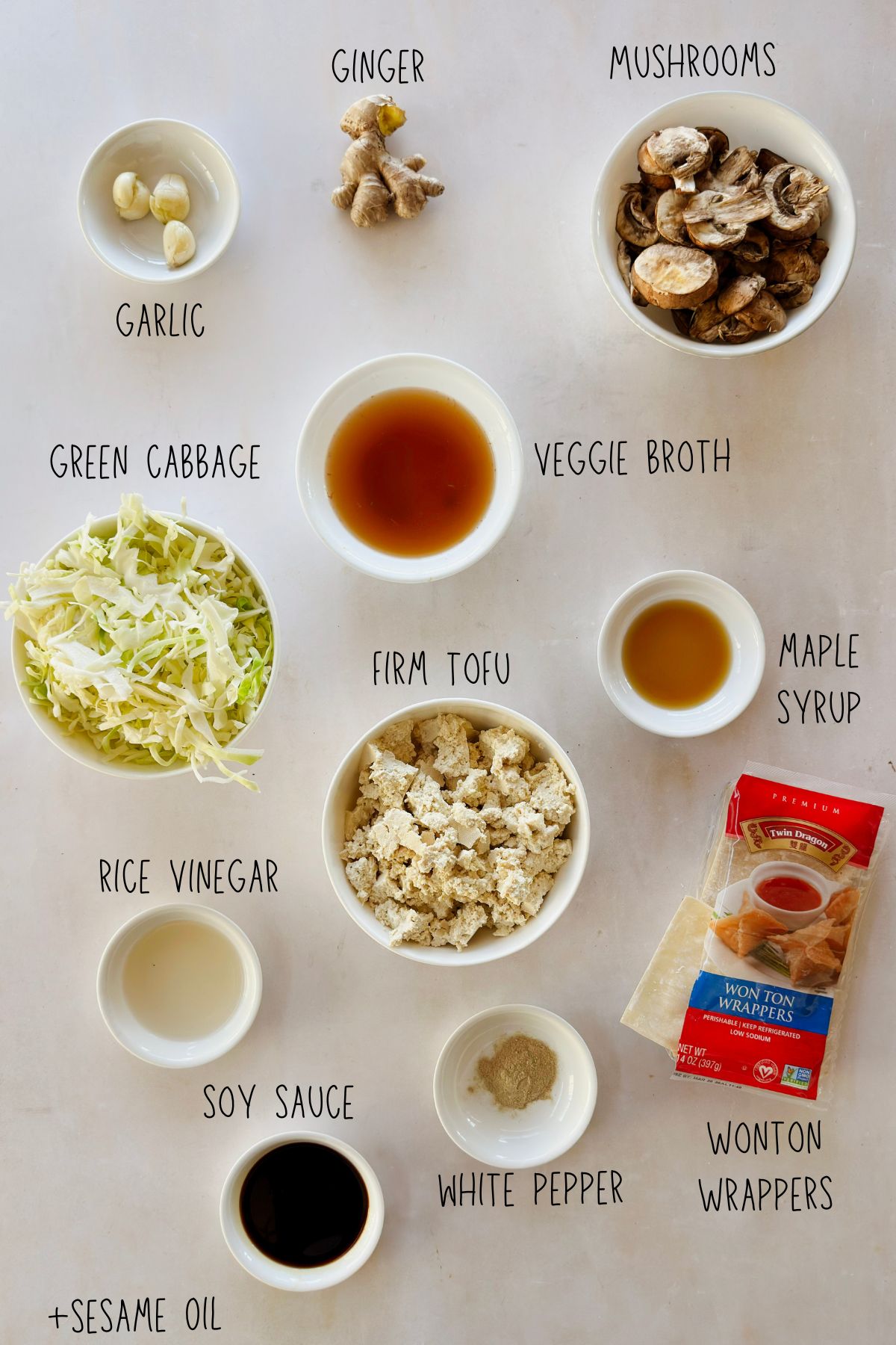 Flat lay of dumpling lasagna ingredients arranged on a light surface with labels. Small white bowls hold garlic cloves, sliced mushrooms, shredded green cabbage, crumbled firm tofu, rice vinegar, soy sauce, white pepper, maple syrup, and vegetable broth. A knob of fresh ginger sits above, and a package of wonton wrappers rests to the side with a few wrappers visible. 