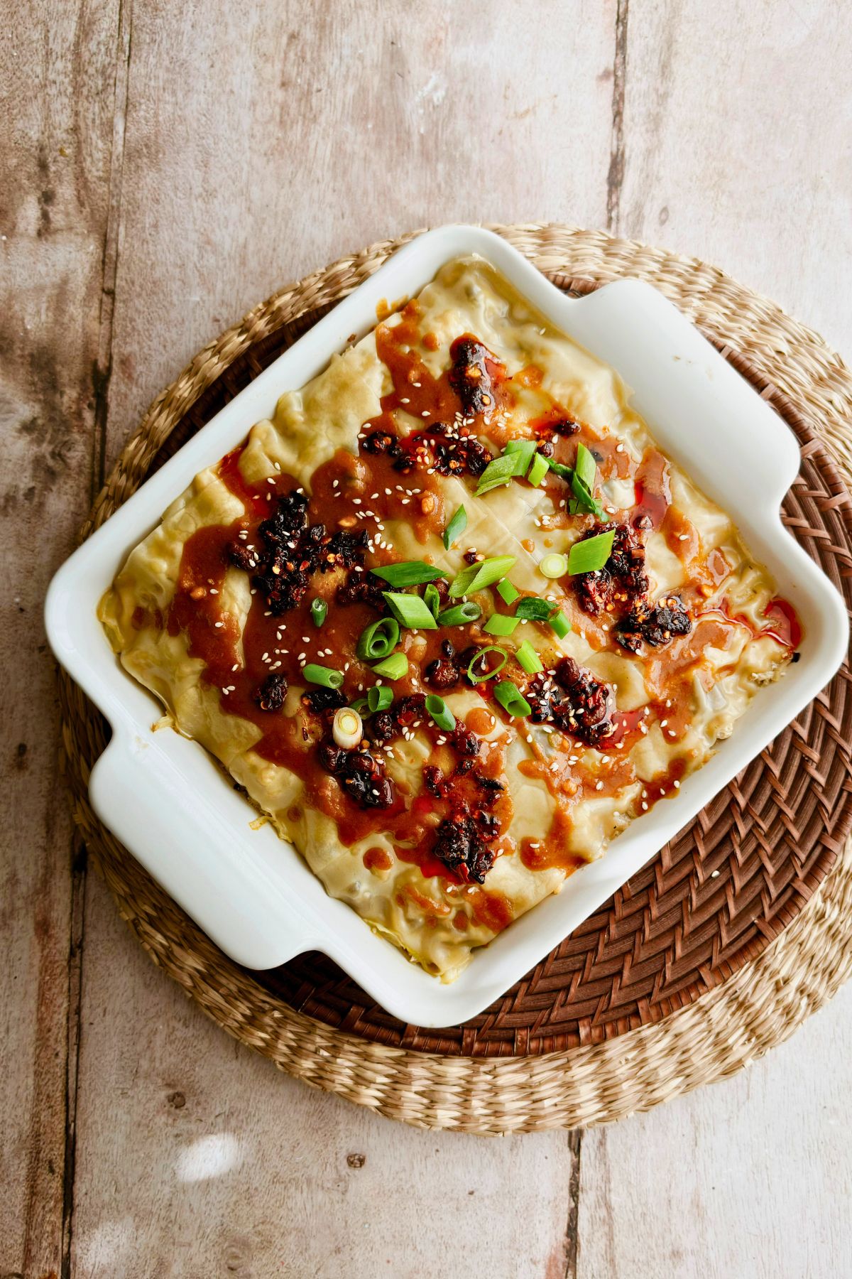 Overhead shot of vegan dumpling lasagna in a white baking dish on a woven trivet, topped with savory sauce, crispy chili, sesame seeds, and fresh green onions.