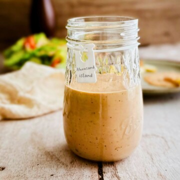 A glass mason jar half filled with creamy pale pink vegan Thousand Island dressing with a small white handwritten label tag reading thousand island, blurred salad and wooden background behind it.