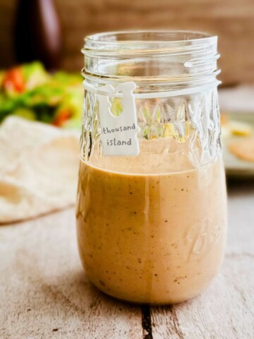 A glass mason jar half filled with creamy pale pink vegan Thousand Island dressing with a small white handwritten label tag reading thousand island, blurred salad and wooden background behind it.