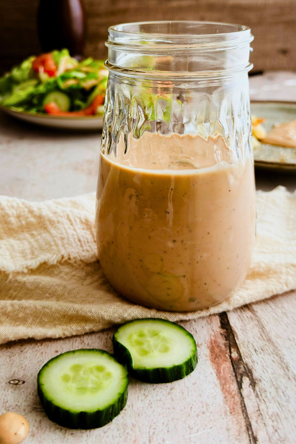 A glass mason jar half filled with creamy pale pink vegan Thousand Island dressing, blurred salad and wooden background behind it.