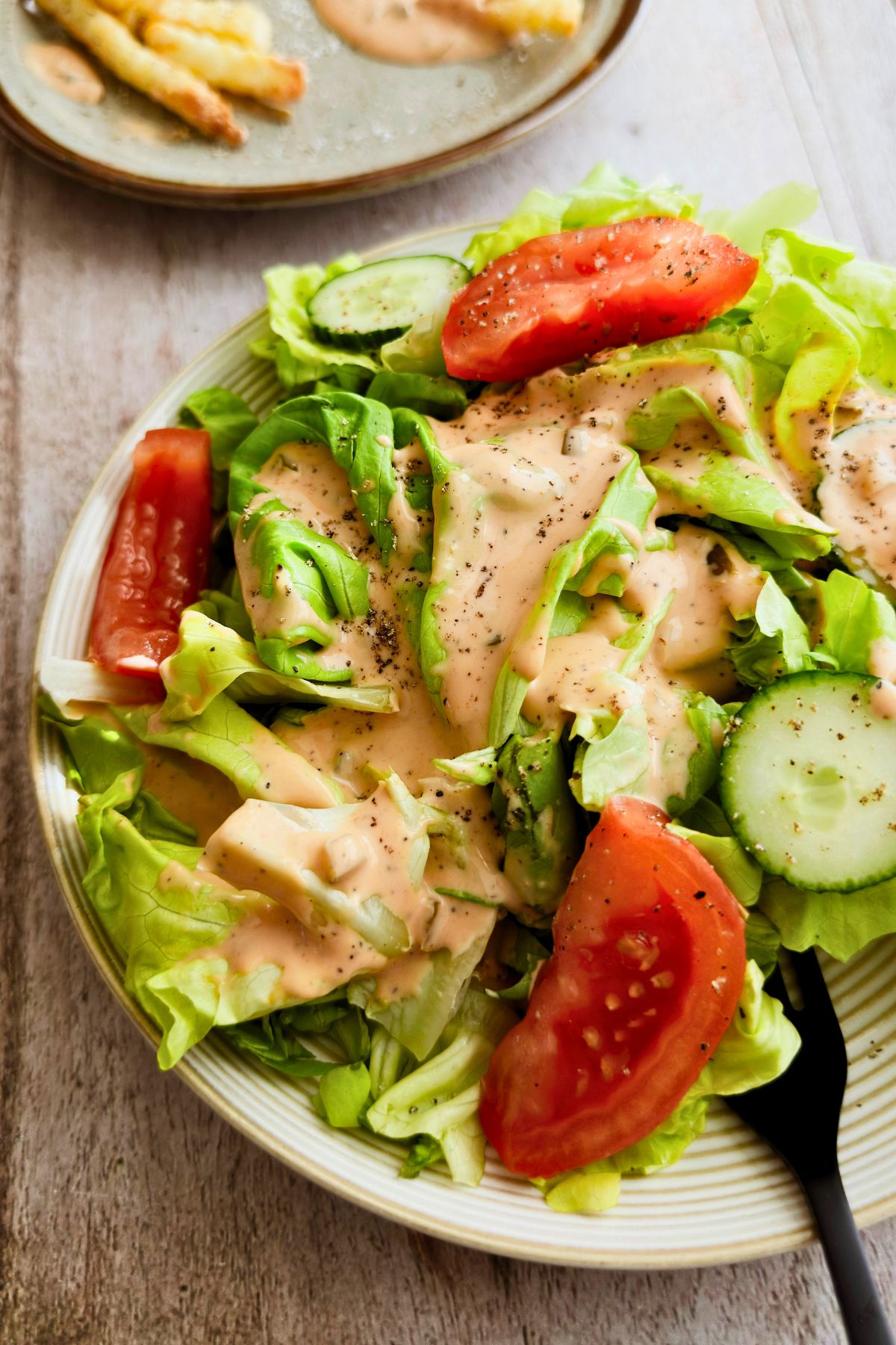 Close up of a butter lettuce salad generously coated in creamy pink-orange vegan Thousand Island dressing with thick tomato wedges and sliced cucumber on a ridged ceramic plate, black fork resting on the side.