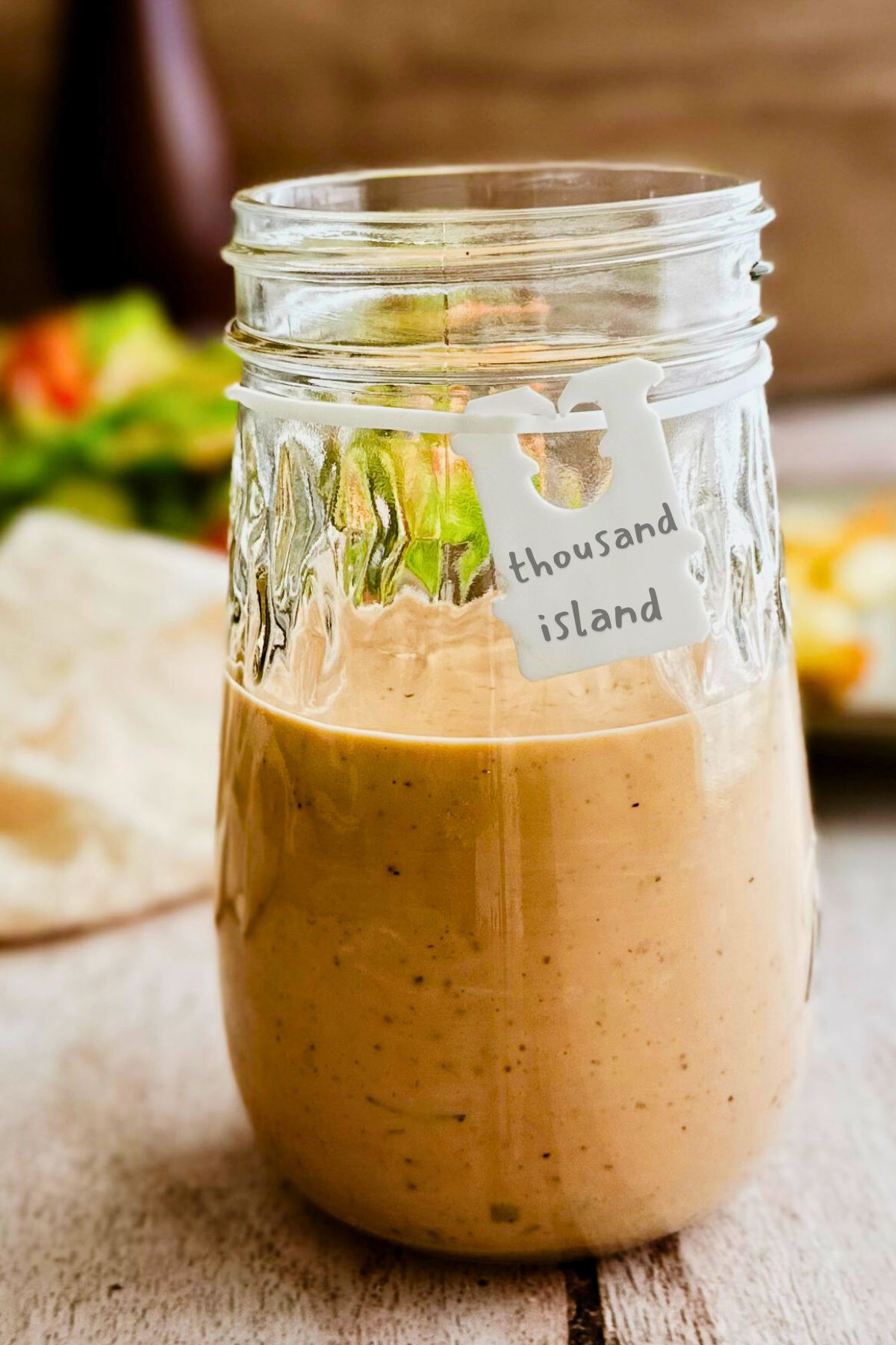 A glass mason jar half filled with creamy pale pink vegan Thousand Island dressing with a small white handwritten label tag reading thousand island, blurred salad and wooden background behind it.