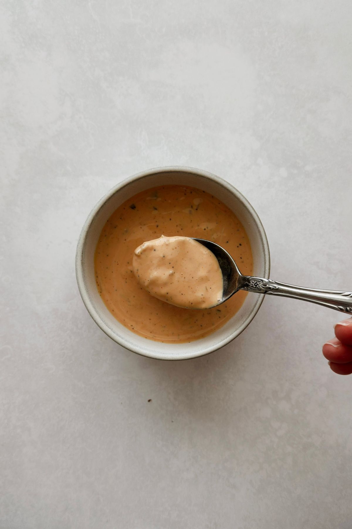 Overhead shot of a rustic white ceramic bowl filled with creamy vegan Thousand Island dressing on a light grey surface, a vintage silver spoon lifting a heaped scoop of dressing with a hand visible at the edge.