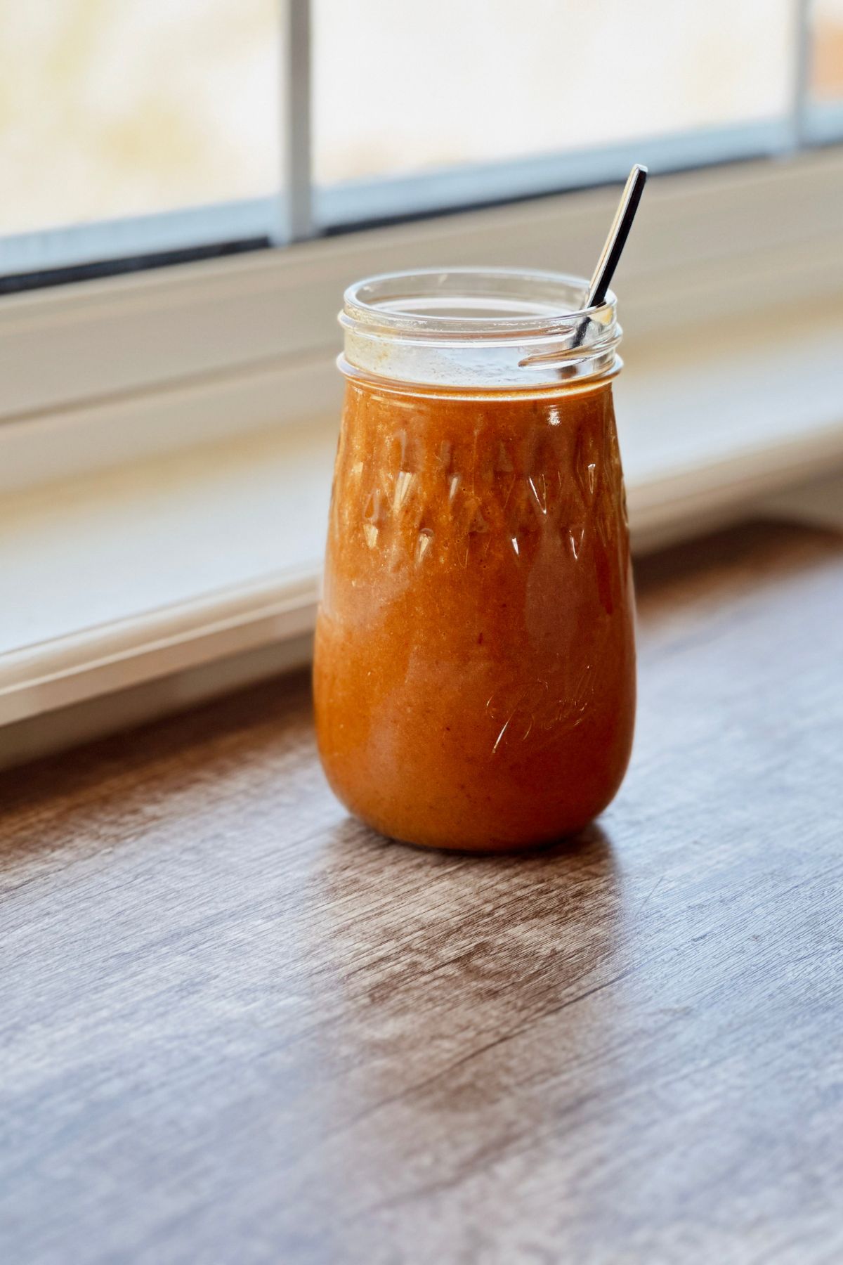 A mason jar filled with smooth, deep orange chipotle vinaigrette with a metal spoon resting inside, sitting on a wooden surface in front of a sunlit window.