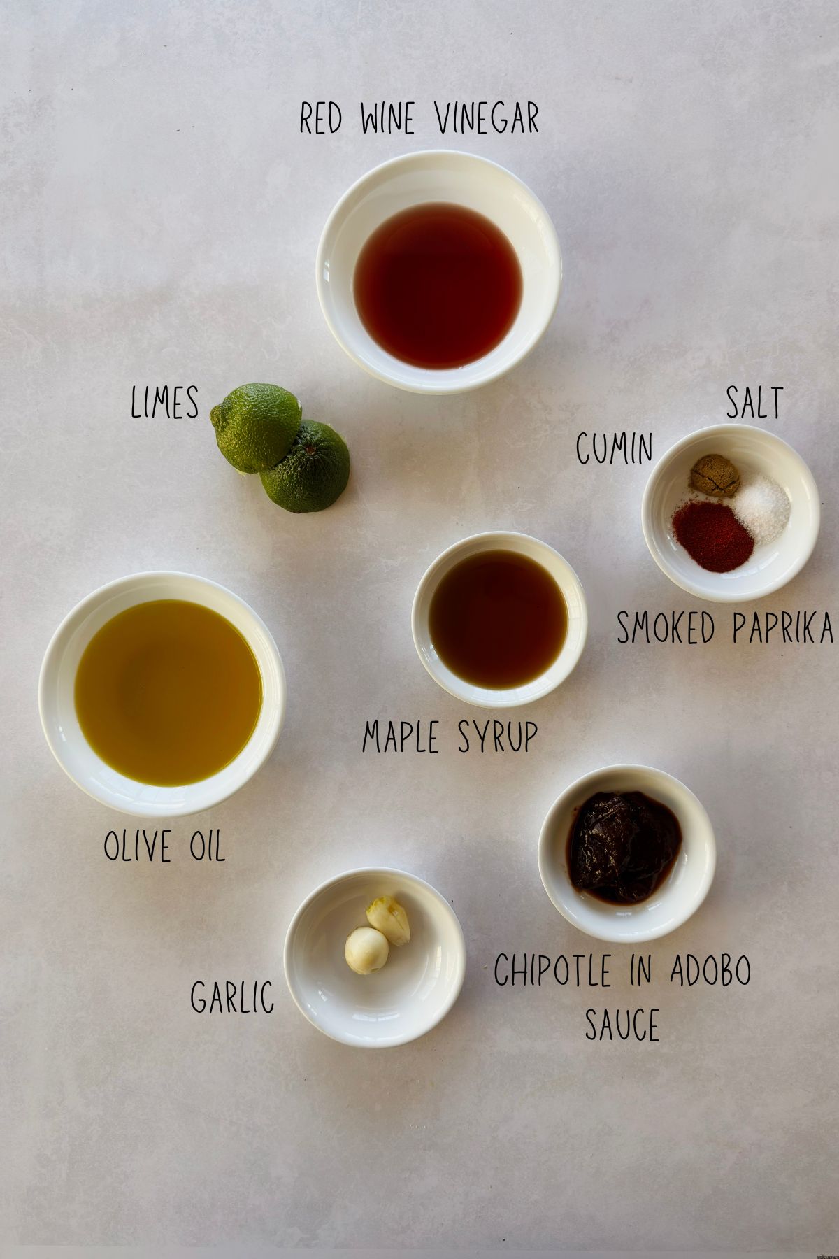Flat lay of chipotle vinaigrette ingredients in small white bowls on a light background, labeled with red wine vinegar, limes, olive oil, maple syrup, garlic, chipotle in adobo sauce, cumin, salt, and smoked paprika.