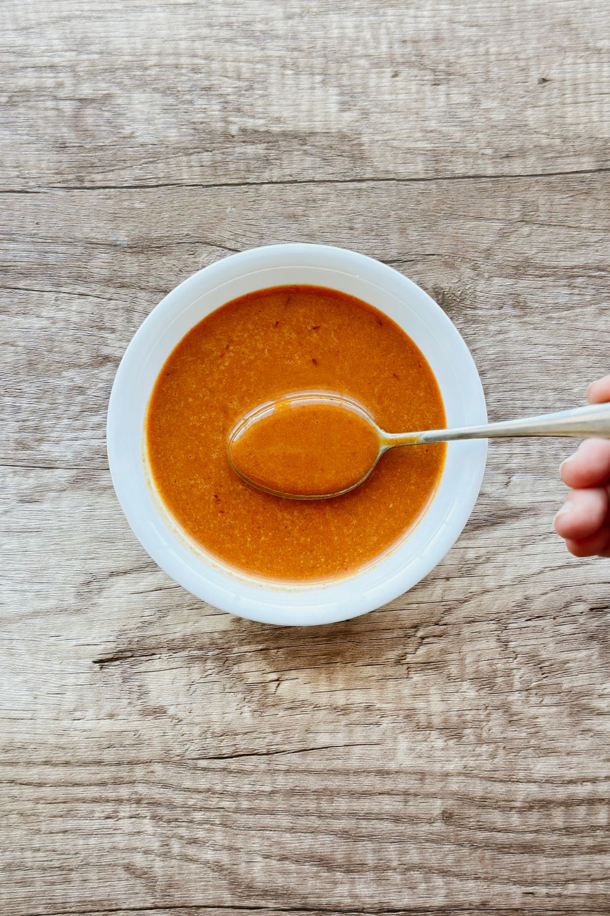 A hand holding a spoon over a white bowl filled with smooth, deep orange chipotle vinaigrette, set on a rustic wood surface.