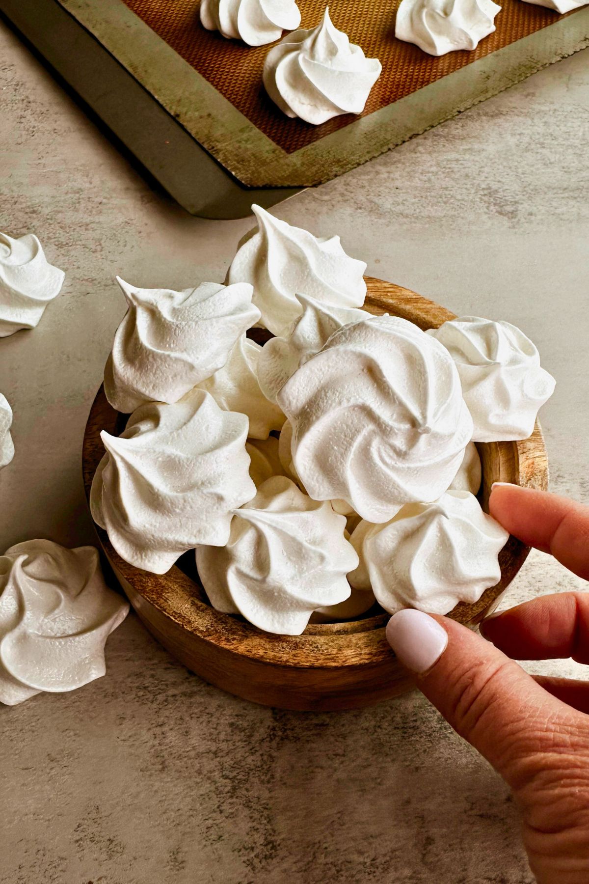A hand reaching toward a round wooden bowl filled with Cuban suspiros meringue cookies on a warm gray surface. Additional suspiros are scattered on the surface to the left, and a silicone-lined baking sheet with more suspiros is visible in the background, showing the full yield of the recipe.