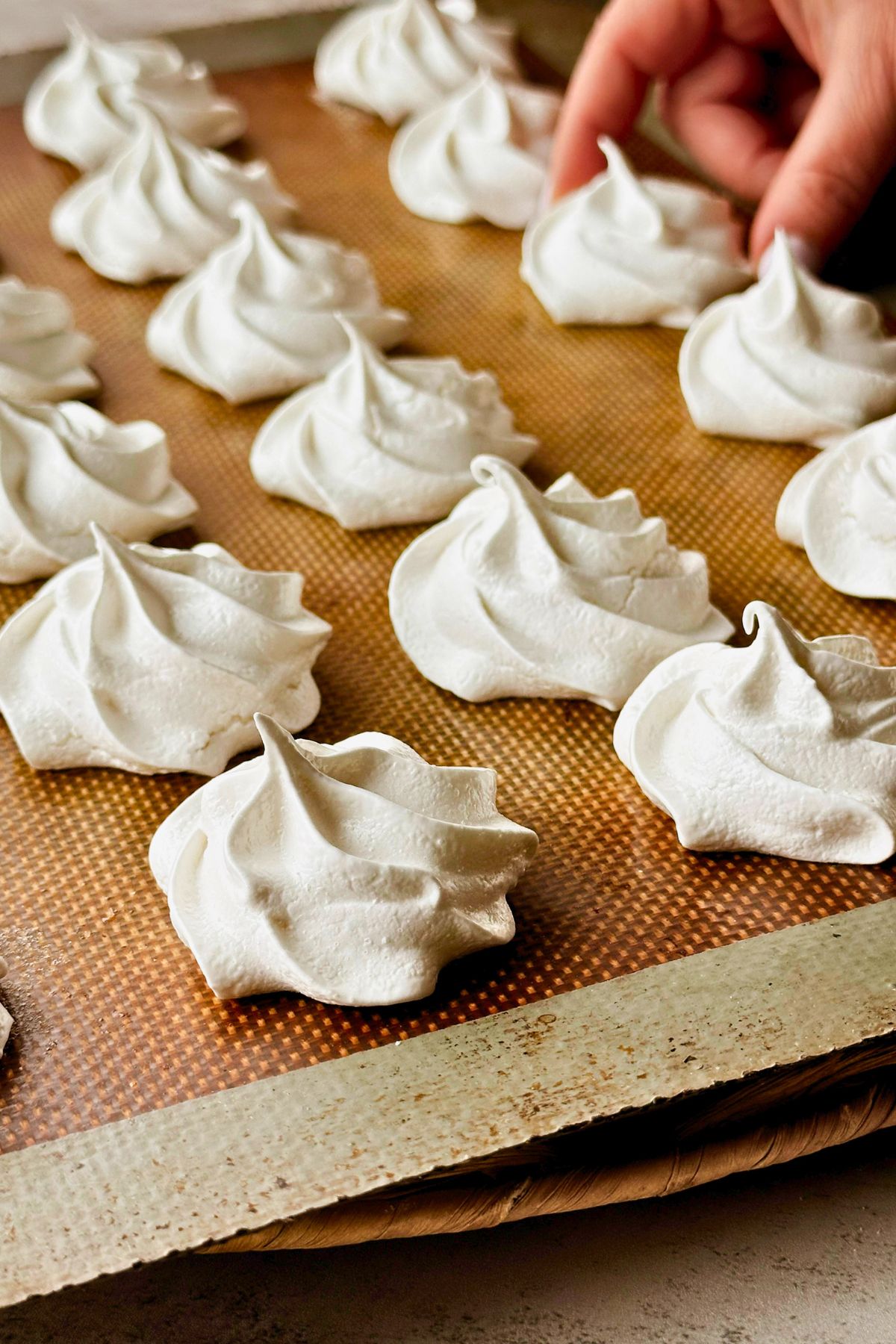 Close-up of freshly baked Cuban suspiros on a silicone-lined baking sheet, showing their crisp white exterior and piped rosette texture in detail. A hand reaches in from the upper right corner to pick one up, giving a sense of their light, airy size.