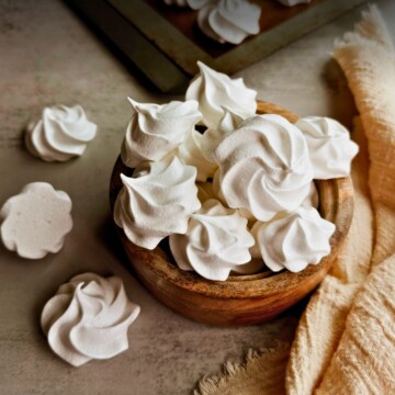 A wooden bowl overflowing with white Cuban suspiros meringue cookies on a warm gray surface, with a golden linen cloth, scattered cookies, and a baking sheet visible in the background.