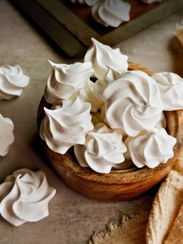 A wooden bowl overflowing with white Cuban suspiros meringue cookies on a warm gray surface, with a golden linen cloth, scattered cookies, and a baking sheet visible in the background.