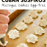 A hand piping glossy white vegan Cuban suspiros meringue rosettes onto a silicone-lined baking sheet, with the text "How to Make Cuban Suspiros Meringue Cookies Egg-Free" overlaid at the top and nutsandtwigs.com in the lower left corner.