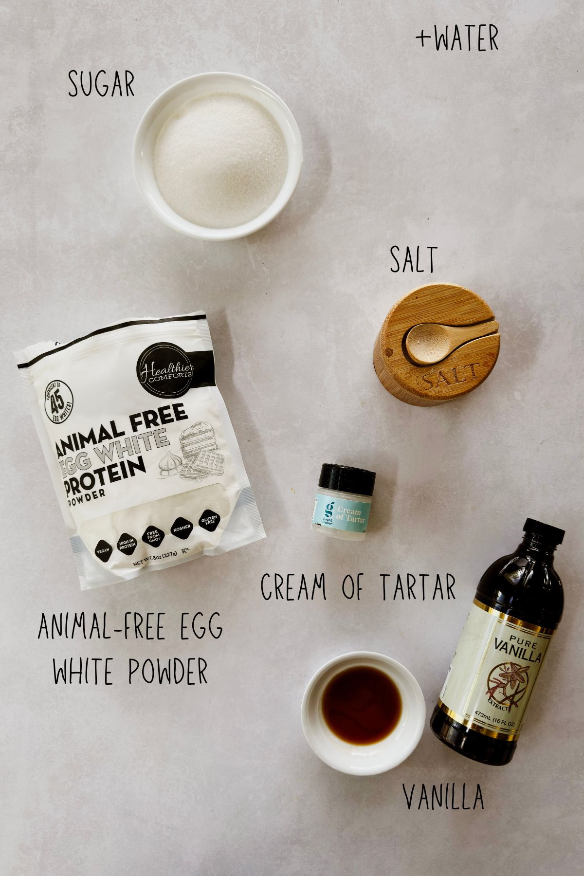 Flat lay of Cuban suspiros ingredients on a light gray surface, labeled: a white bowl of granulated sugar, a wooden salt cellar, a small jar of cream of tartar, a small white bowl of vanilla extract next to a bottle of pure vanilla extract, and a bag of Healthier Comforts Animal-Free Egg White Protein Powder. A "+water" label appears in the upper right corner indicating water is also needed but not shown.